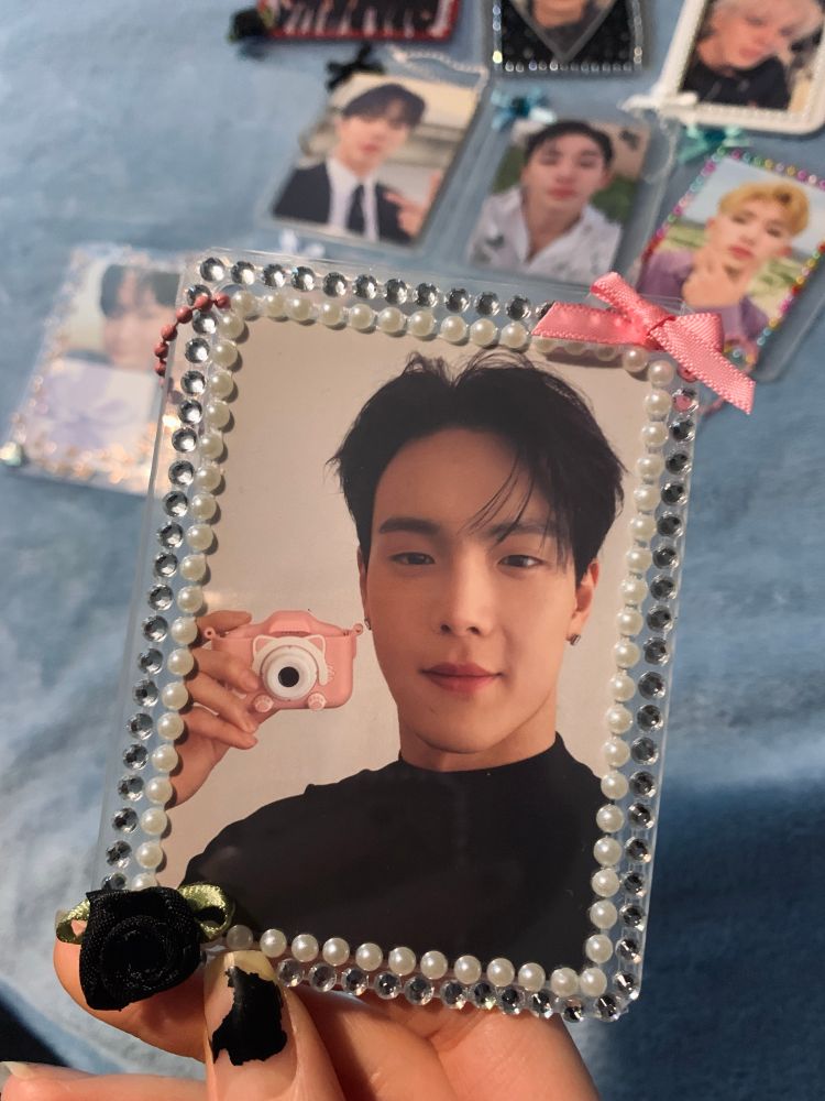 A photocard of monsta x shownu inside a hard photocard sleeve with plastic white and silver jewels with a black rose and light pink  bow on it 