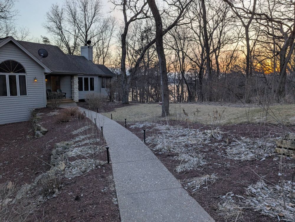 A small blue house is to the left, a lot of trees to the right and between the bare winter trees, you can see a frozen lake as the last rays of sunlight fade at sunset