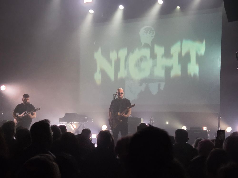 Photo hyper flou avec un écran dans le fond et un "night" super flou. Les deux musiciens sur scène guitares en main, un des deux le chanteur porte des lunettes noires. On voit les têtes du public. Lumière légère et brume relative