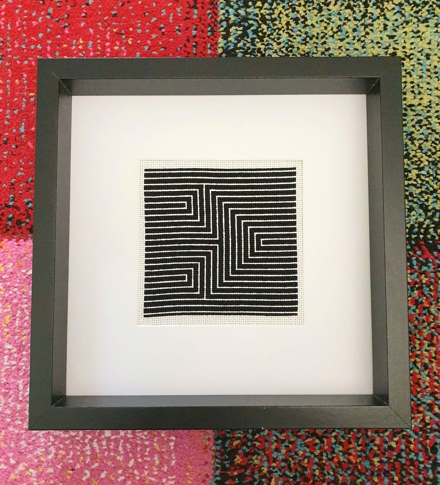 Das Bild zeigt ein gerahmtes Kreuzstich-Stickbild in Schwarz-Weiß. Das Muster besteht aus zwei geometrischen Pfeilformen, die sich spiegeln und aus eng nebeneinander verlaufenden weißen Linien auf schwarzem Hintergrund gebildet werden. Das Design wirkt sehr modern und optisch ansprechend, fast hypnotisch. Der Rahmen ist schlicht, schwarz und quadratisch, das Stickbild ist mittig auf weißem Passepartout montiert. Der Hintergrund besteht aus einem bunten, mehrfarbigen Teppich in Rot, Pink, Blau und Grün.