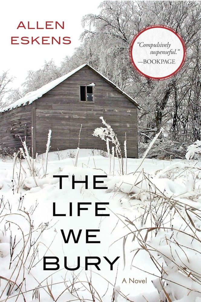 The cover of the novel The Life We Bury shows a winter foreground with solitary grasses sticking up from snow covered ground and a weathered building, no windows, in the background.