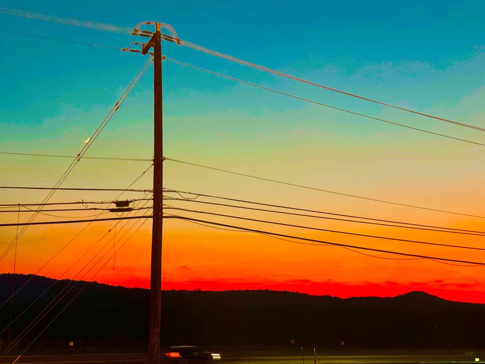 A photo taken near a road, with a blurry car passing near the bottom. The car’s headlights illuminate a small portion of the road. The background is composed of hills cast into shadow and the sky has clear bands of blue at the top, yellow in the middle, fading into orange and a deep red above the hills. There is a telephone pole in the foreground, and a crescent moon appears to be resting on one of the wires that goes from the top of the pole to the ground at a 45-degree angle. 