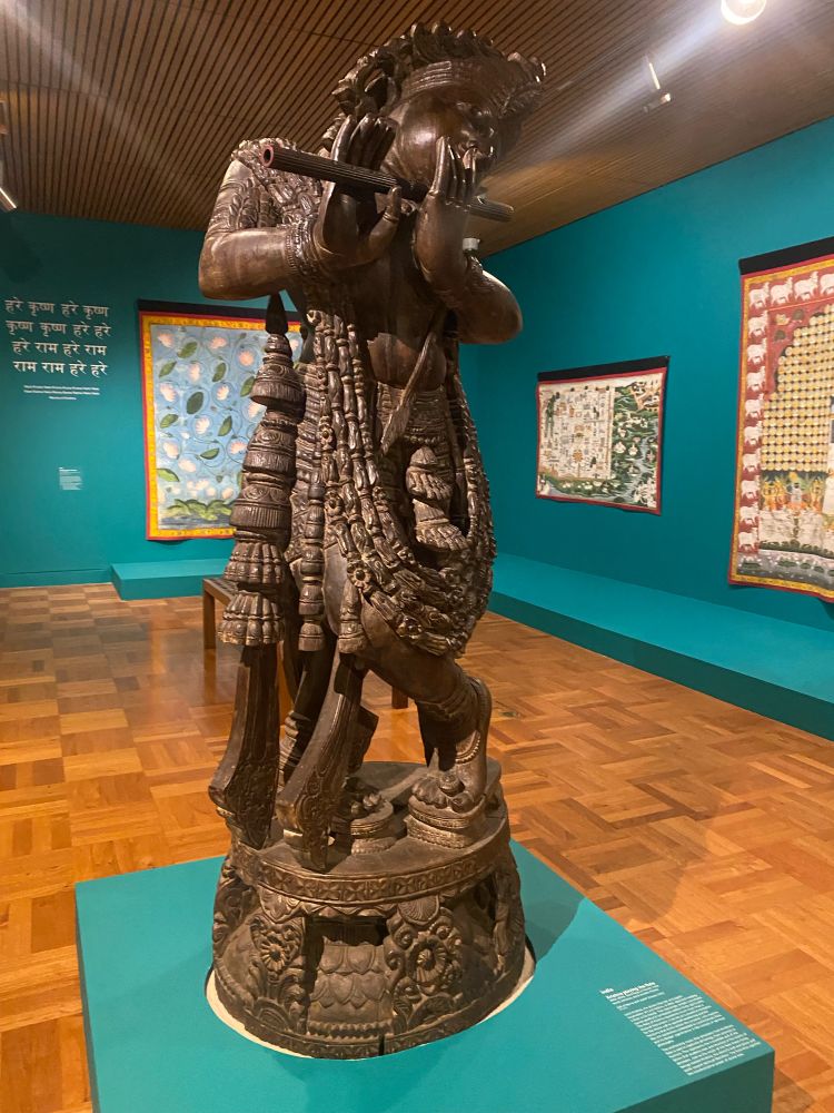 A sculpture: Krishna Playing a Flute, Art Gallery of South Australia. It’s life-sized, dark wood, ornate, standing in on an azure block in the middle of the gallery floor. There are other Indian artworks in the background 