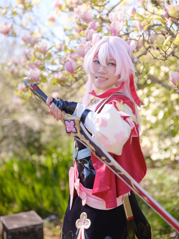 a photo of connie cosplaying March 7th from Honkai: Star Rail in her Hunt Form. connie is holding one sword across their body while facing to the left and looking to the right. a magnolia tree with pink flowers is blooming in the background. 