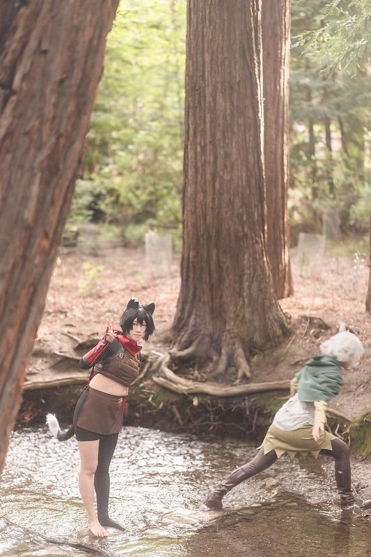 a photo of connie cosplaying Izutsumi. they are standing in a river making a peace sign with their right hand. Mithrun is to their right, walking across with river. they are slightly blurry due to motion blur. the river is surrounded by redwood trees.