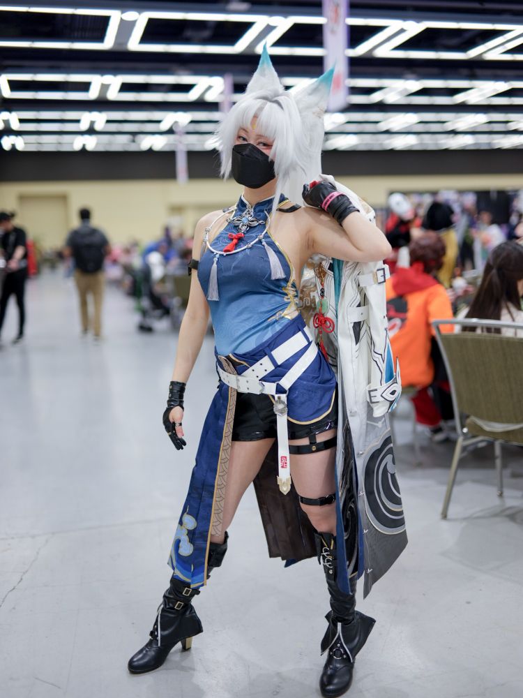 a photo of connie cosplaying Feixiao. they are standing in the artist alley with Feixao's jacket thrown over one shoulder. they are wearing a black KF94 respirator.