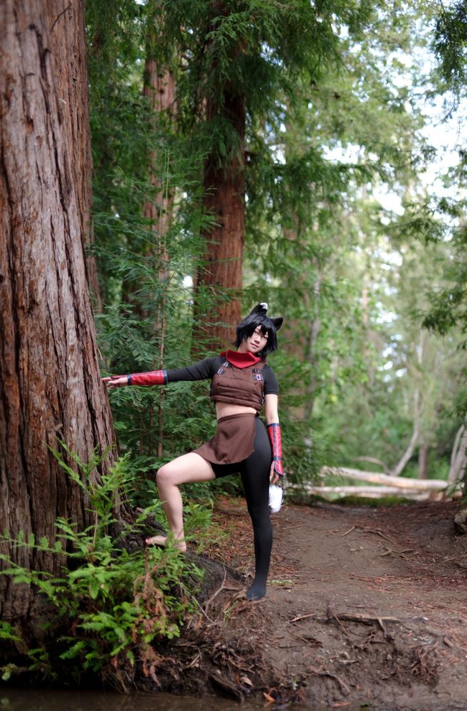 a photo of connie cosplaying Izutsumi from Dungeon Meshi. they are standing on a dirt path while facing the camera with one hand touching a large redwood tree. more green trees can be seen in the background.