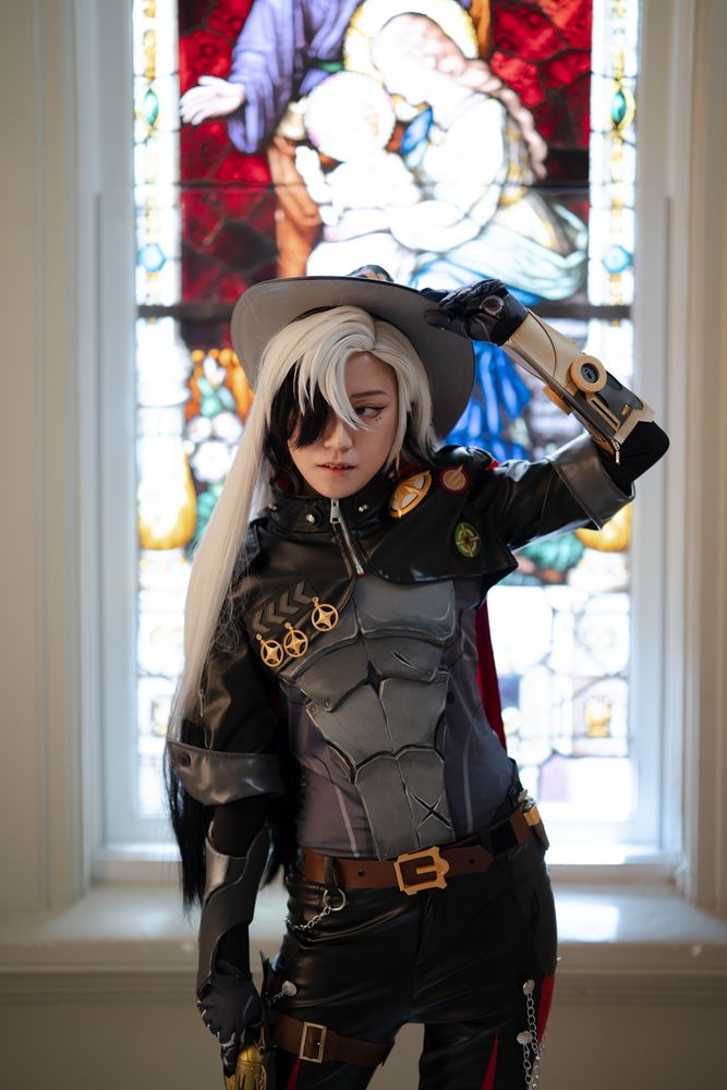 a halfbody photo of connie cosplaying Boothill from Honkai: Star Rail. They are standing in front of a colorful stained glass window and looking to the side while adjusting their cowboy hat.