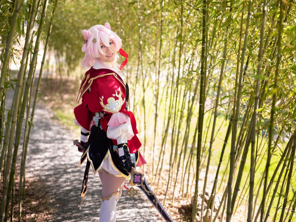 a photo of connie cosplaying March 7th from Honkai: Star Rail in her Hunt Form. connie is standing with their back partially to the viewer while holding two swords. they are smiling and looking back while standing on a path lined with bamboo plants. 