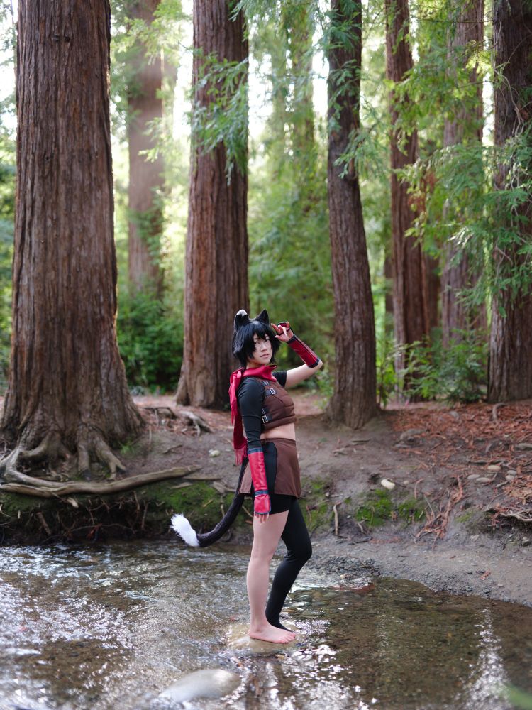 a photo of connie cosplaying Izutsumi from Dungeon Meshi. they are standing in a river with tall redwood trees behind them. they are looking into the camera with one hand shielding their eyes from the sun.