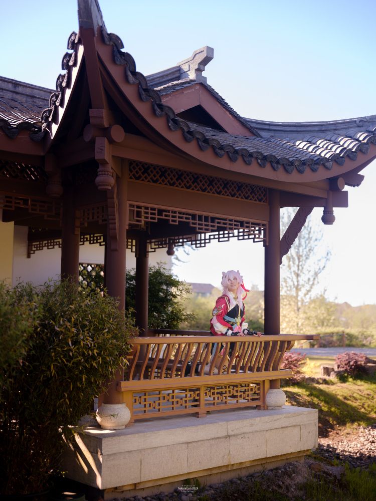 a photo of connie cosplaying March 7th from Honkai: Star Rail in her Hunt form. connie is standing in a pavilion while leaning over a wooden railing while smiling and looking past the camera. the shot is taken from far away and the entire pavilion can be seen. 