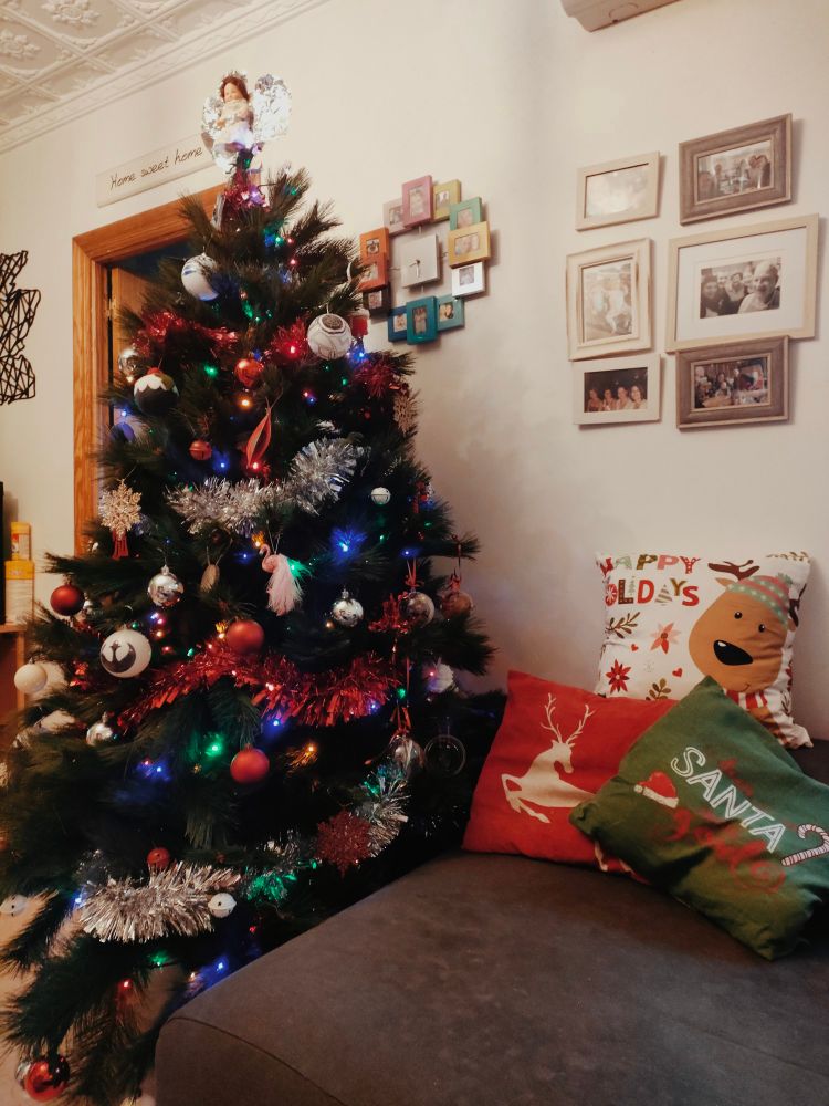 A tall Christmas tree decorated with red and gold, with a vintage dolly angel on top stands beside a comfortable sofa with festive cushions.