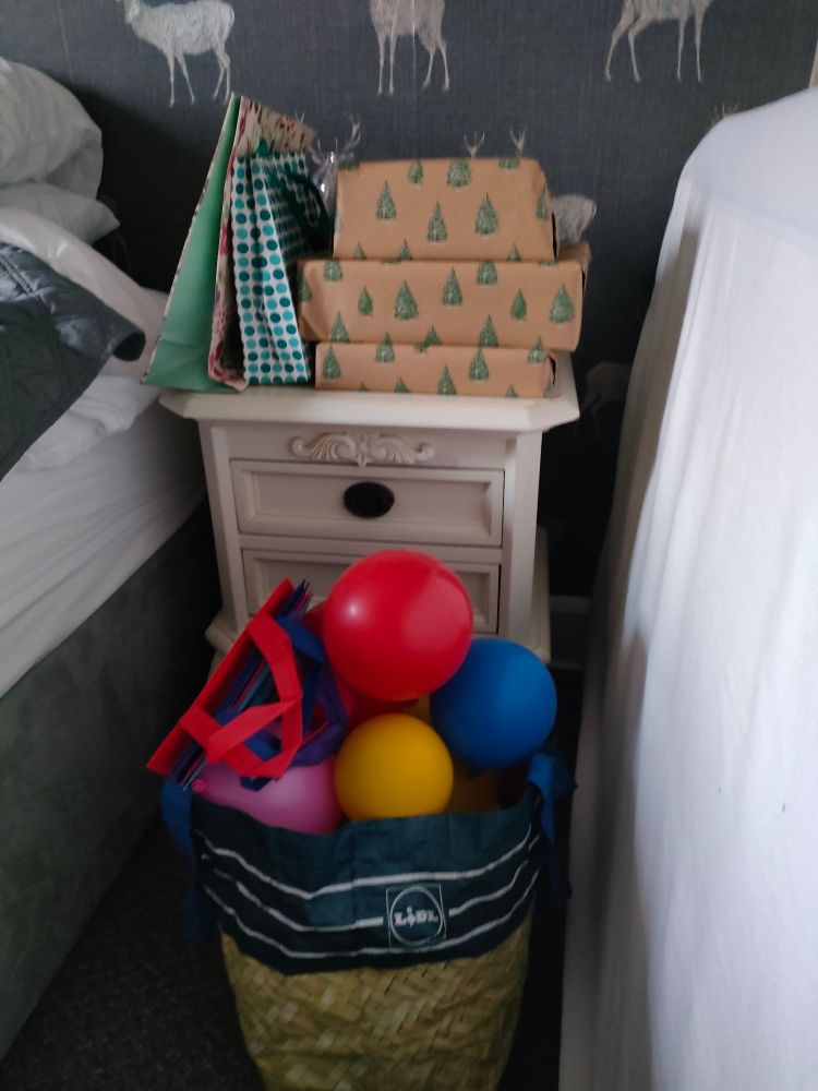 A pile of Christmas presents sits atop a white bedside table in a cozy bedroom. A shopping bag sits on the floor in front, filled with Christmas goodies, glow sticks and balloons spilling over the top.