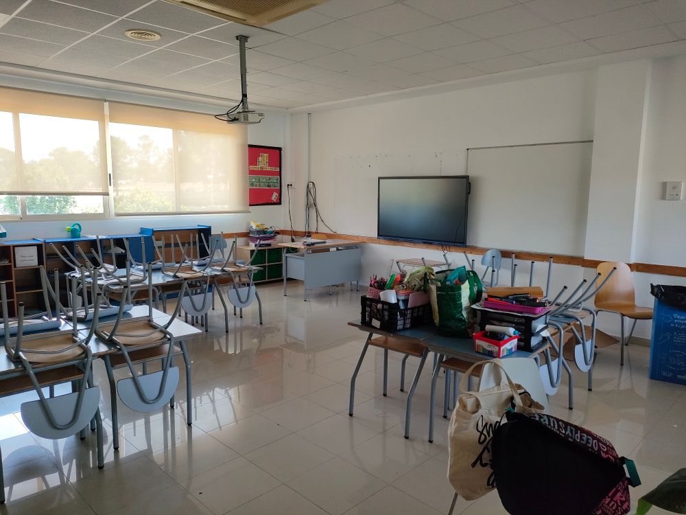 An empty classroom with chairs up on desks. Some book cases and storage units are visible around the room, and an interactive board hangs on the wall beside a whiteboard.