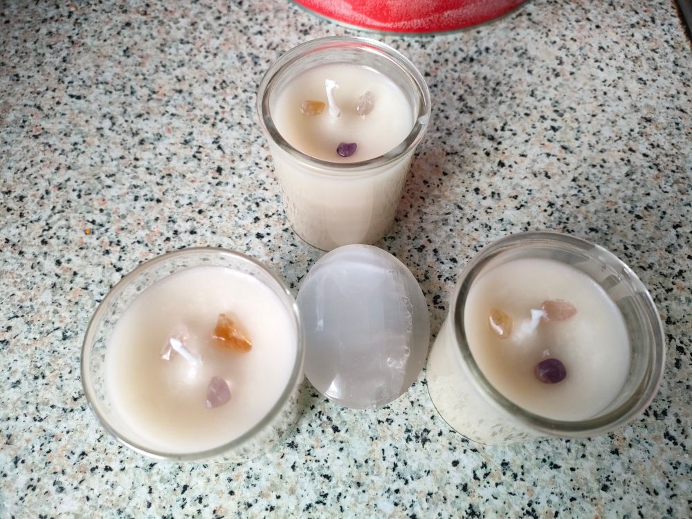 Three white homemade recycled candles in glass jars surround a white gemstone. Each candle is topped with a purple amethyst chip, a yellow citrine chip and a pale pink rose quartz chip.