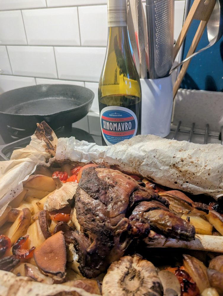 In the foreground, a fish of kleftiko. Behind that is a frying pan, a jar of kitchen utensils and a bottle of xinomavro wine from Macedonia. 