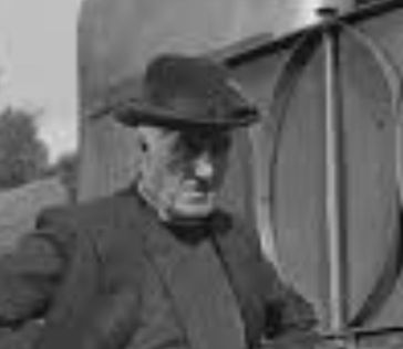 A black and white still from The Titchfield Thunderbolt showing the Bishop standing next  to a train. He is wearing a black hat with a medium high crown . The brim is attached to the crown with some sort of tape or ribbon, but this is not very noticeable in this image. 