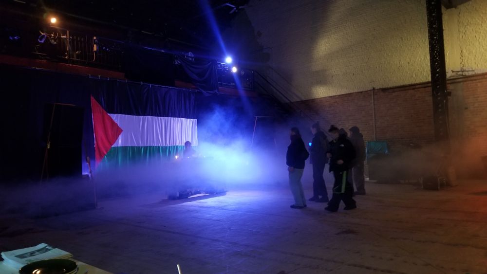 Photo d'une grande pièce de spectacle avec une dj devant un énorme drapeau palestinien et un petit groupe qui danse devant. Une machine à fumée donne une ambiance éthérée 