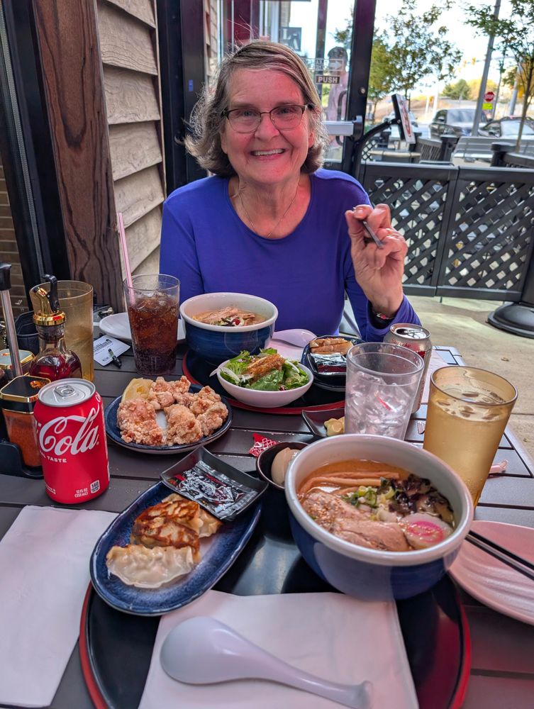 A woman sitting at an outdoor table at a Japanese restaurant. In front of her is karaage, two bowls of ramen, gyoza and two cans of soda.