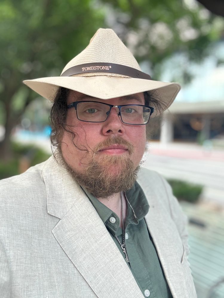 A bearded, four-eyed doofus is wearing beat-up Panama hat, a cream colored linen jacket, and a green linen shirt. He’s also wearing a Thor’s Hammer and his hair is not cooperating.
