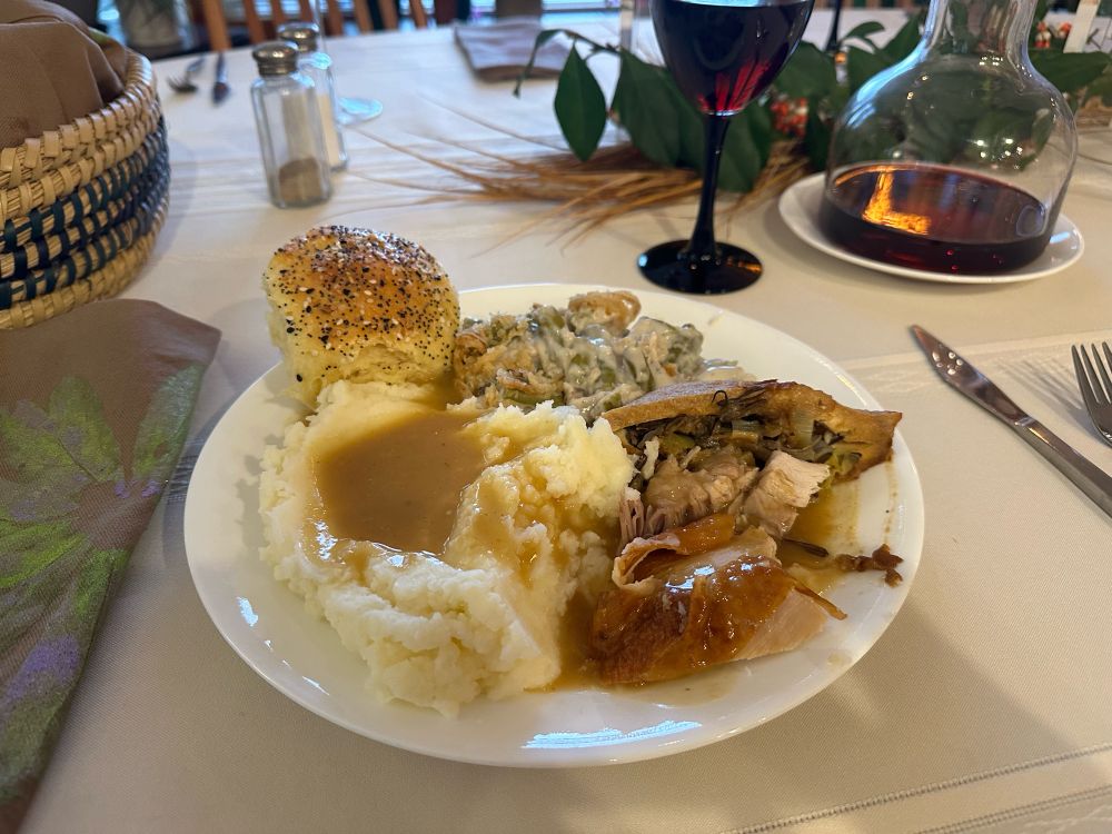 Dinner plate with turkey, mashed potatoes, green bean casserole, an everything roll, wild-rice-stuffed tofu triangle, and gravy, with a glass of red wine and other table settings in the background