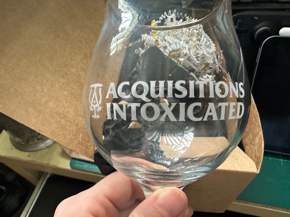 The same glass has been emptied of paper and is being held by the photographer's left hand for better visibility of the 'ACQUISITIONS INTOXICATED' text and logo, which resembles an A and an I overlaid on each other, with curves extending up from the crossbar of the A to form the tulip shape of the beer glass.