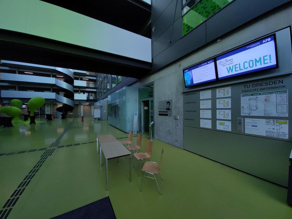 View into a university hallway, with chairs, tables, a staircase, and digital signage saying "RIOT Summit September 3 - 5, 2025" / "Meet in Dresden!" / "Welcome"