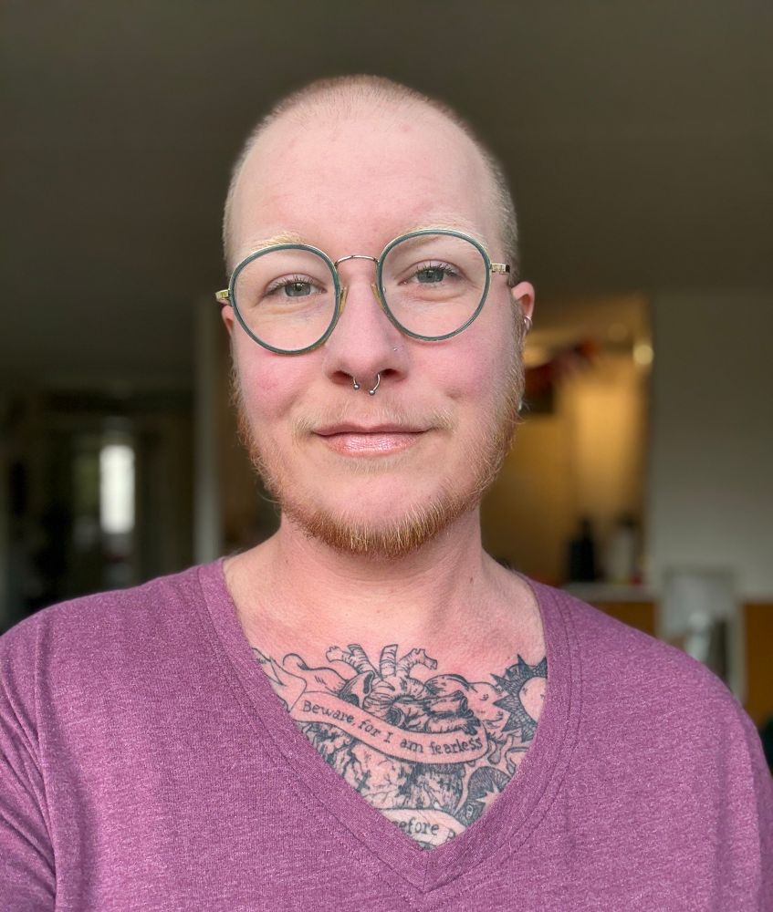 een selfie van Jan, een witte non-binaire persoon met gemillimeterd haar, een bril en een baardje. hen heeft geblondeerde wenkbrauwen en een septum piercing. hen draagt een auberginekleurig shirt met v-hals waardoor hun borst tattoo gedeeltelijk zichtbaar is.
