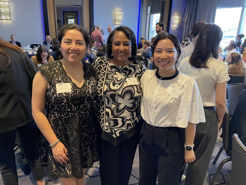Three women standing together smiling at a networking event with blue accent lighting. The woman on the left wears a black lace dress, the woman in the center wears a black and white patterned dress with geometric floral designs, and the woman on the right wears a white blouse with black trim and black pants.