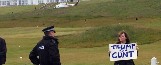 An image of Janey Godley at Turnberry Golf Course holding the famous sign "Trump is a cunt" in front of a police officer. The club mansion in the background, with a helicopter parked.

Image from her blog post "I welcomed Trump to Scotland". https://janeygodley.com/2016/06/28/welcomed-trump-scotland .