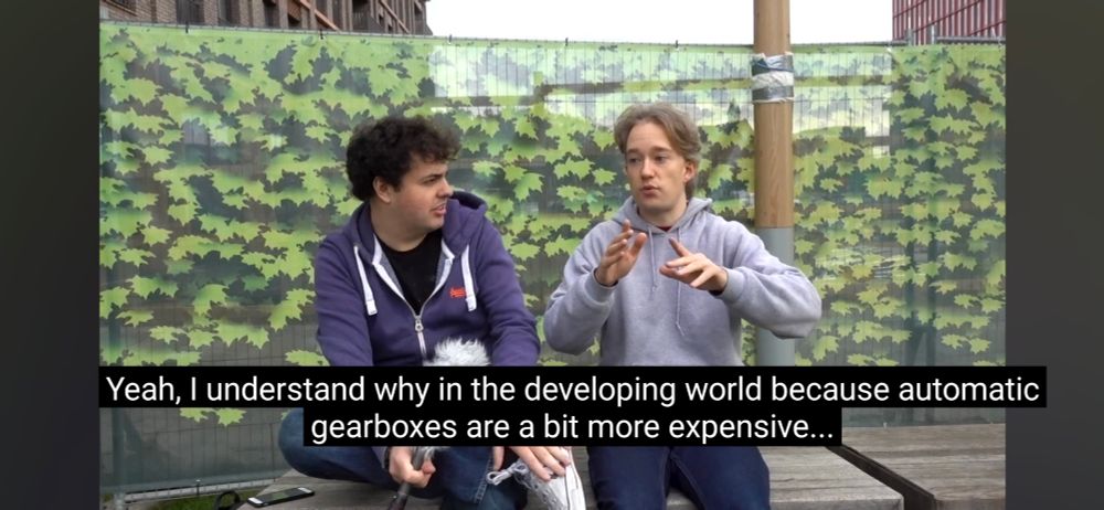 screenshot of the "Matt and Toms Controversial opinions" youtube video. matt and tom are sitting on a bench in front of a green tarp, arguing over whether or not card should have automatic gearboxes.