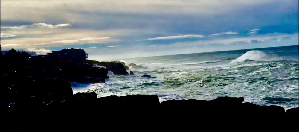 Photo is of a early spring day at the coast. There is blue sky over the ocean with coastal haze and clouds over land. The ocean is turquoise with foamy white waves as it approaches  rocky shores.