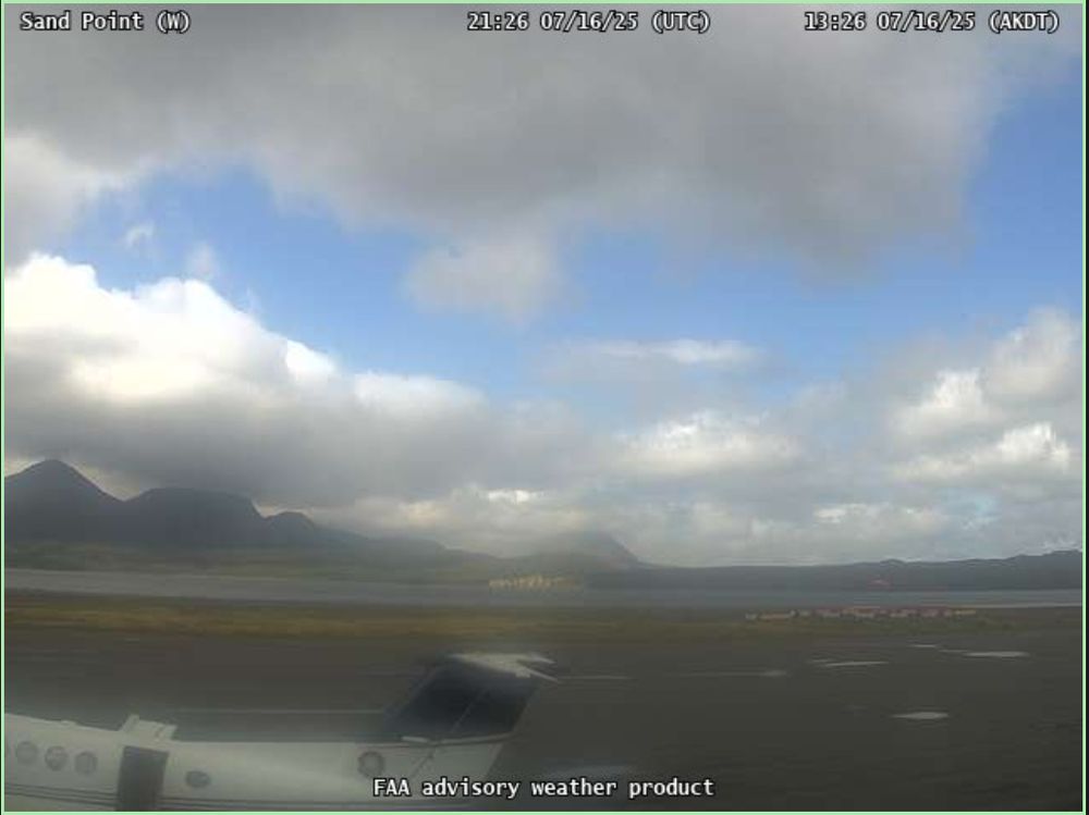 A fuzzy weathercam image of a small passenger jet sitting on a tarmac runway, with clouds overhead and some blue sky. Mountains and the ocean are visible in the background.