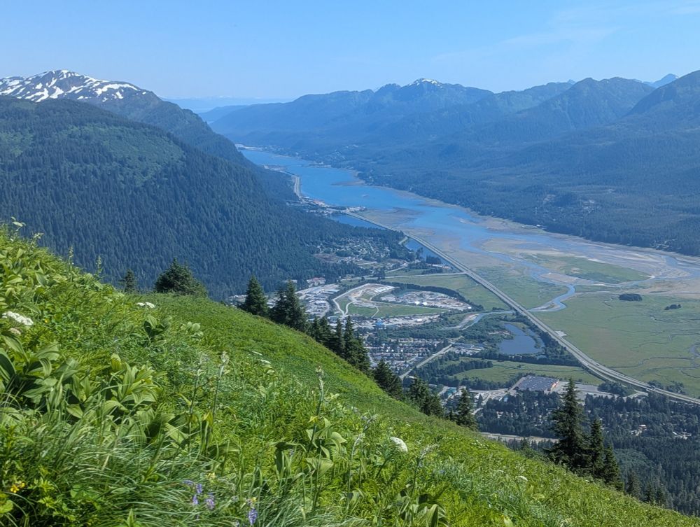 A blue sky and green landscape view of Juneau, Alaska neighborhood Lemon Creek from a trail on Thunder Mountain. Nestled in mountains, a wetlands transitioning into ocean sit on one side of a road with a small neighborhood and landfill on the other side.