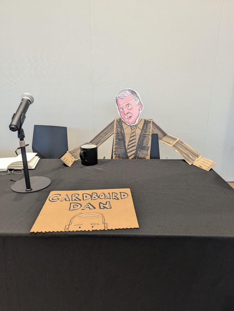 A cartoon cardboard cutout of Senator Dan Sullivan sits at a black desk, with a sign that says "Cardboard Dan" in front of him. There's a black coffee cup near his hand as well as a microphone on the table.