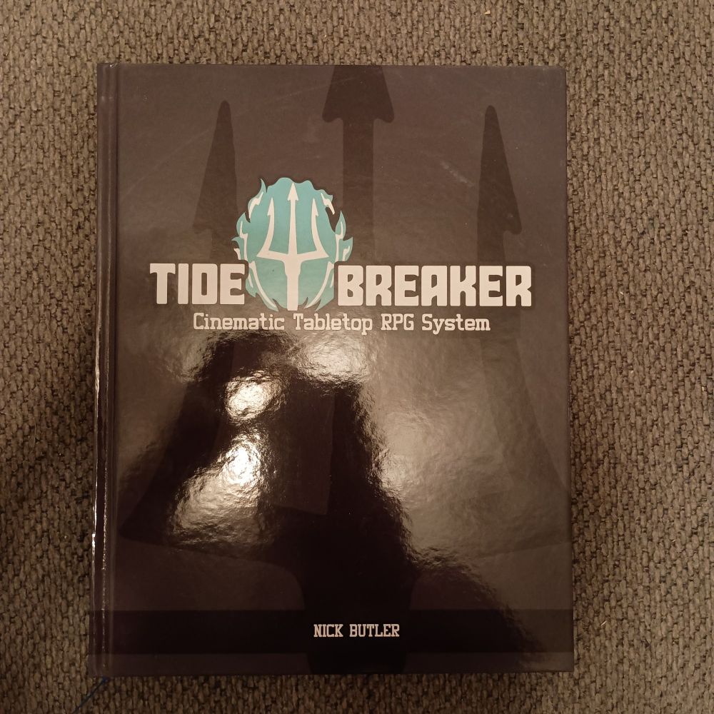 Cover of Tide Breakers. Black cover with the title in white and  the head of a trident (also white) surrounded by blue. My shadow is also clearly visible on the cover.