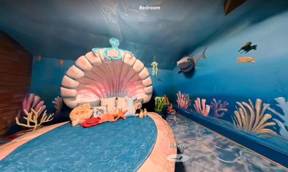 Blue room with a mural of ocean life like coral and fish with a 3-d shark head mounted on the wall. The bed is oval with a fuzzy blue cover and the headboard is a 3-D big, thick, curved, pink scallop shell. There are stuffed critters on the bed like starfish and a teddy bear wearing a scuba mask. The carpet is wibbly blue like looking into a fish tank. 