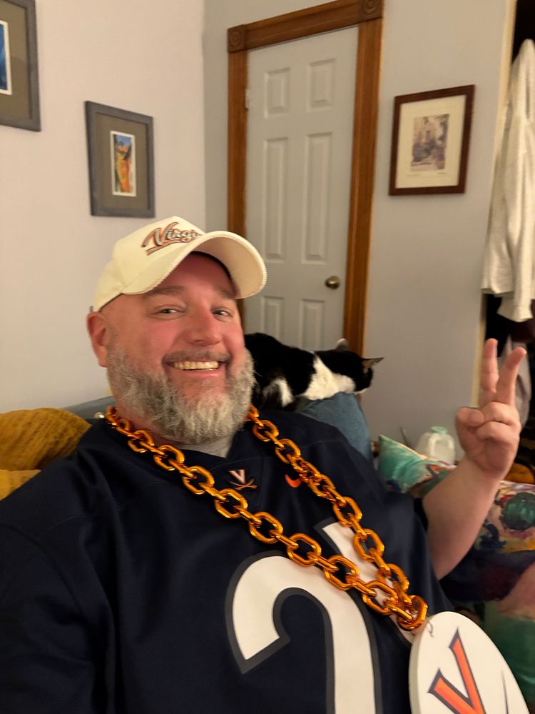 Moderately good looking man with a majestic beard sits next to a gorgeous cat, he’s wearing a chain with a V and sabres on it