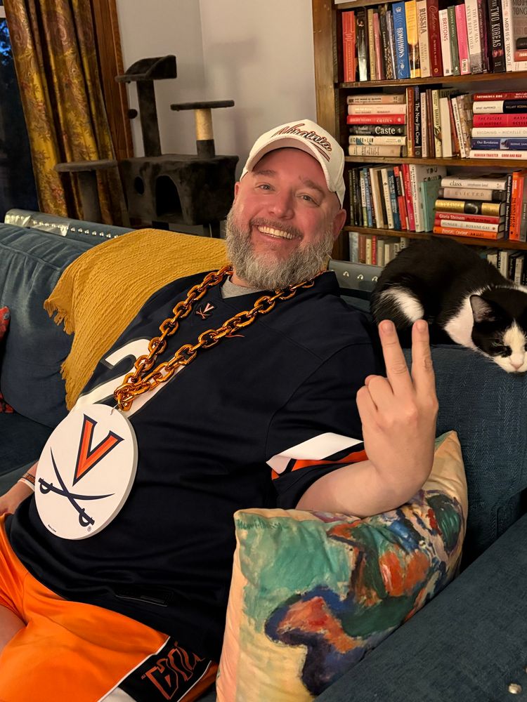 Moderately good looking man with a majestic beard sits next to a gorgeous cat, he’s wearing a chain with a V and sabres on it