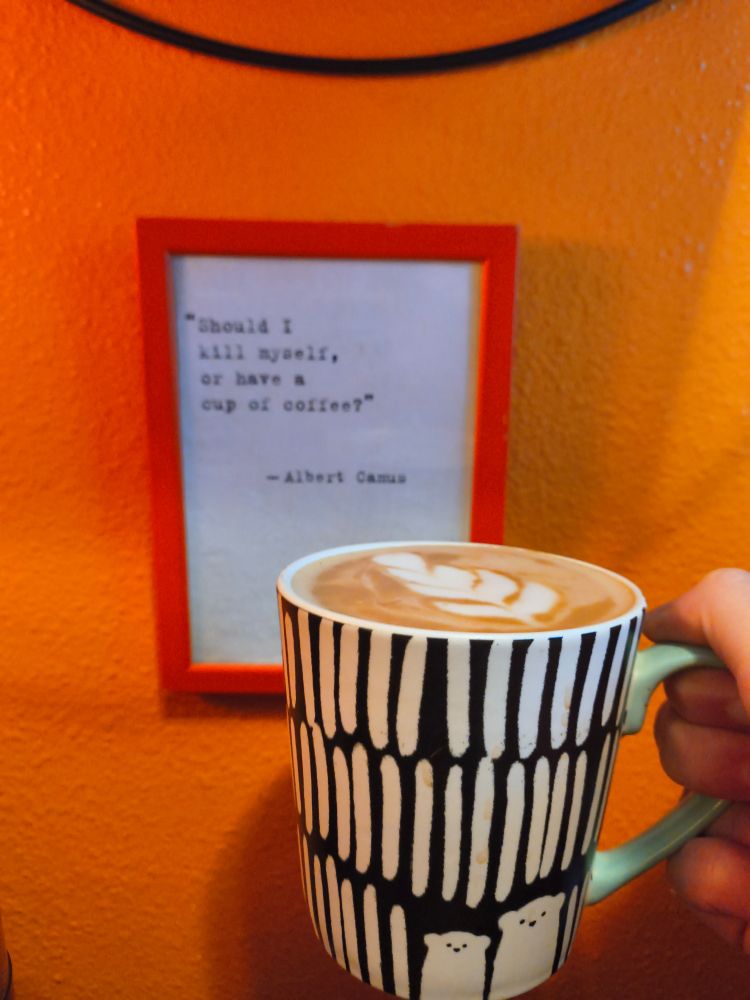 Oat milk latte in a black and white mug with a green handle being held up by a hand. In the background is a framed print of a quote mistakenly cited to Albert Campus that states "Should I kill myself, or have a cup of coffee?". The print is on a white background and in a red frame. It is hanging on an orange wall. 