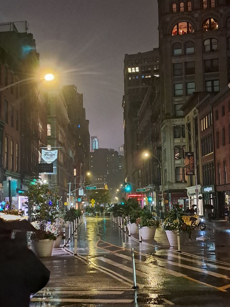 same stretch of Broadway in NYC at night and sopping wet. someone with an umbrella walks past the camera. 