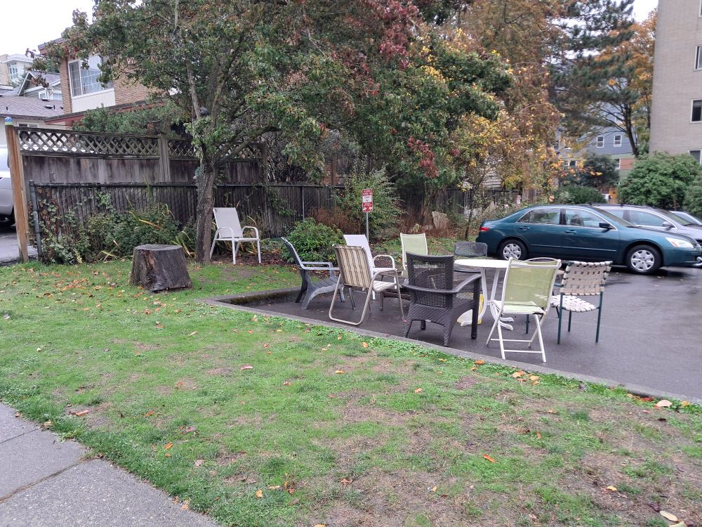 plastic lawn chairs gathered around a small table in a former parking spot. another chair sits beneath a large tree and close to a table sized stump. 