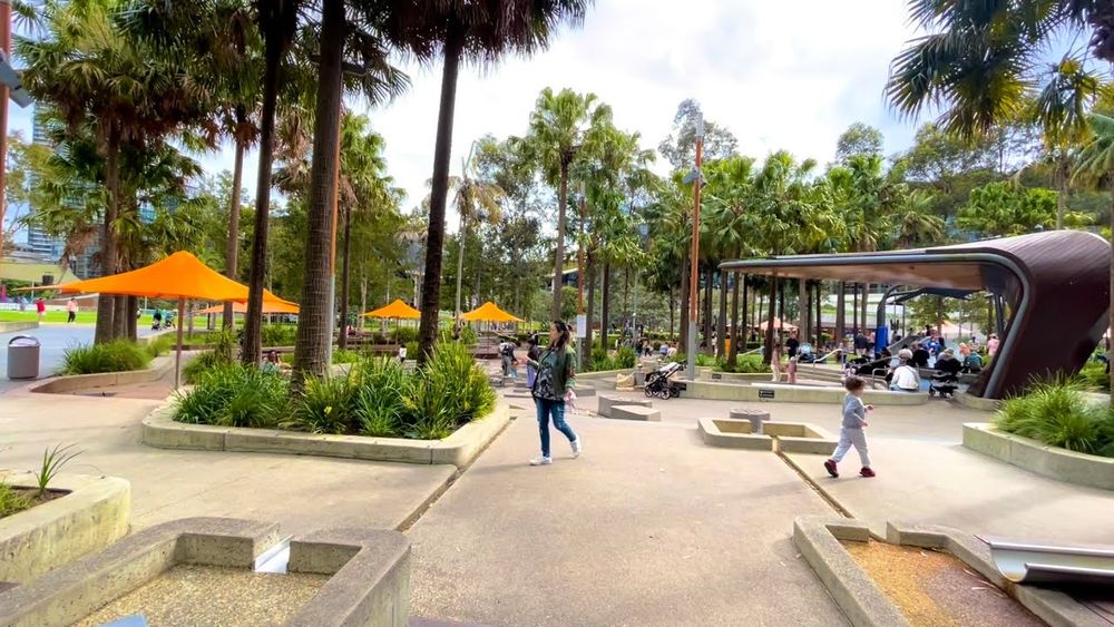 Public square with abundance of large trees, umbrellas, and a modern, abstract-design curved cover, about 5 meters high, offering a bit of shade to seating or a play structure underneath.