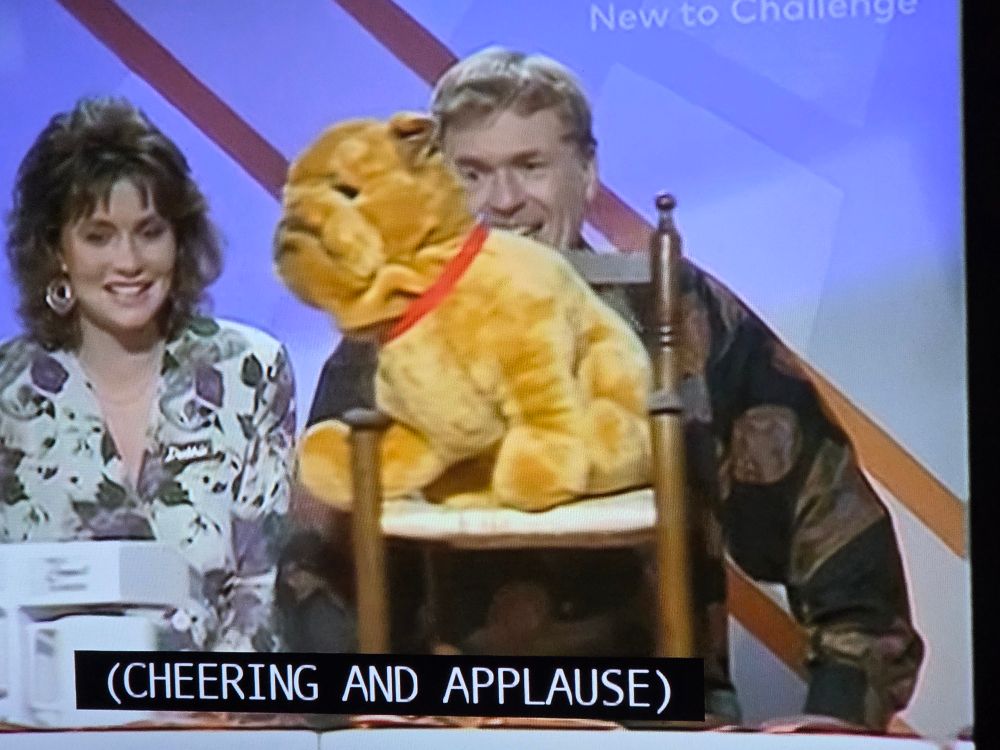 A cuddly toy on the Generation Game conveyor belt watched by two contestants and a subtitle that says CHEERING AND APPLAUSE 