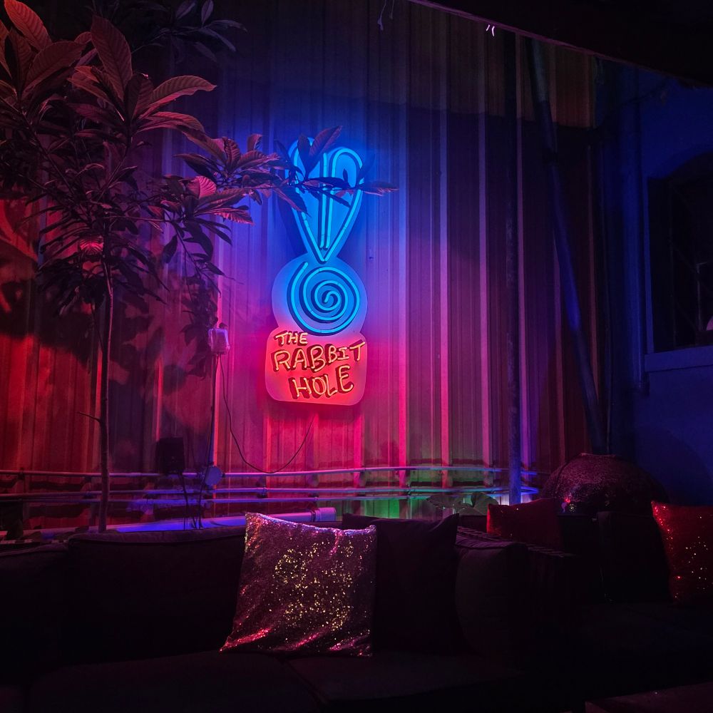 A photo of a neon sign that says "The Rabbit Hole" with bunny ears and a swirl. The lights are blue on top and red on the bottom. Underneath the sign is a couch with sparkly pillows