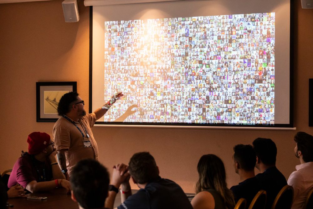 Foto tirada do fundo de uma sala com uma apresentação onde se ve pessoas a frente observando um slide com as fotos de todas as pessoas que estiveram no evento virtual da python Brasil