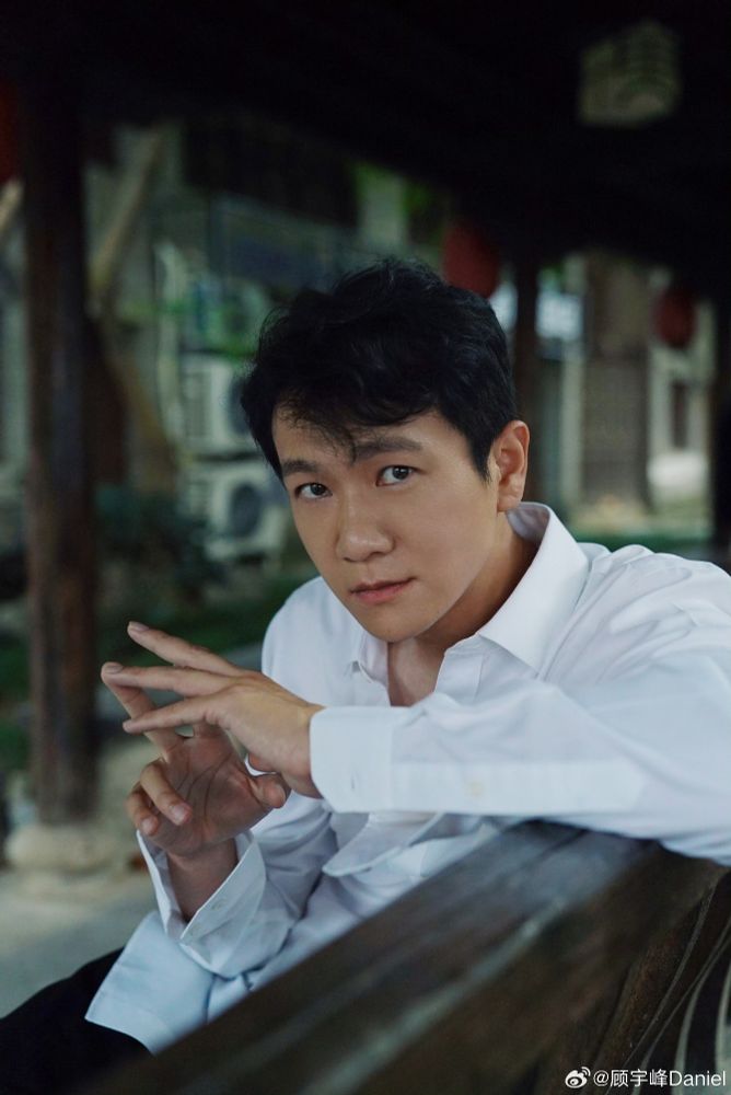 Chinese actor Gu Yufeng, seated on a wooden bench in an outdoor suburban/city area. He wears a white button-up, has his fingers touching each other in a playful way, and is looking at the camera attentively.