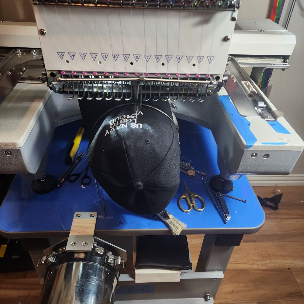 Embroidery machine sewing the phrase "US NAVY LGBTQ PROUD VETERAN" on a black baseball hat.