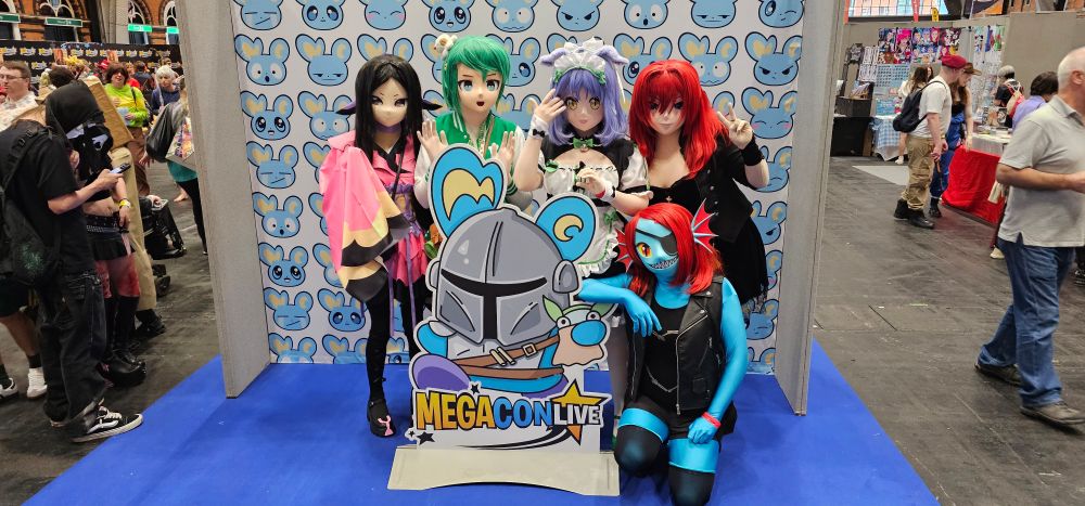 A photo of 5 kigurumi performers, stood around a standee of a blue mascot wearing a knights helmet, and in front of a white background patterned with the face of the blue mascot.