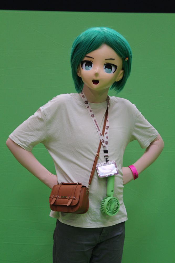 A photograph of a green haired kigurumi character wearing a white shirt and blue chinos in front of a greenscreen. They are looking up slightly with their hands on their hips. 